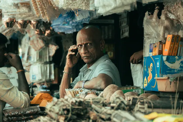 A shopkeeper standing in a busy Indian market, looking towards the camera.